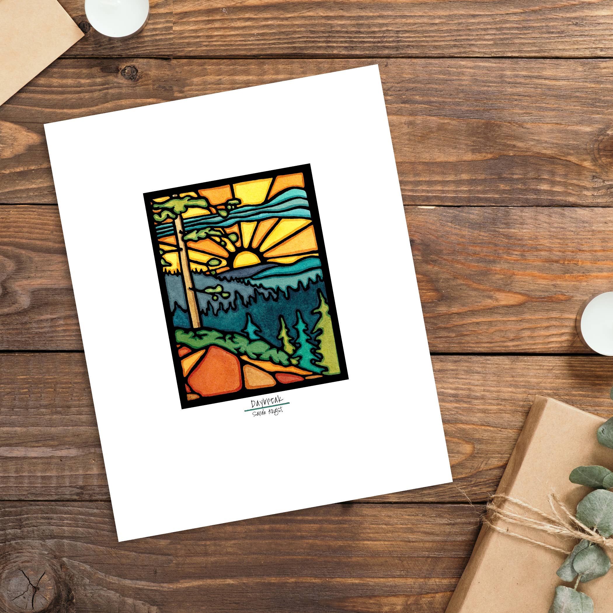 The Artwork: Daybreak Print 11"x14" by Sarah Angst features a stained glass-style sunrise over mountains, trees, and a path. Set with candles, an envelope, and a package, this print adds warmth to any space.