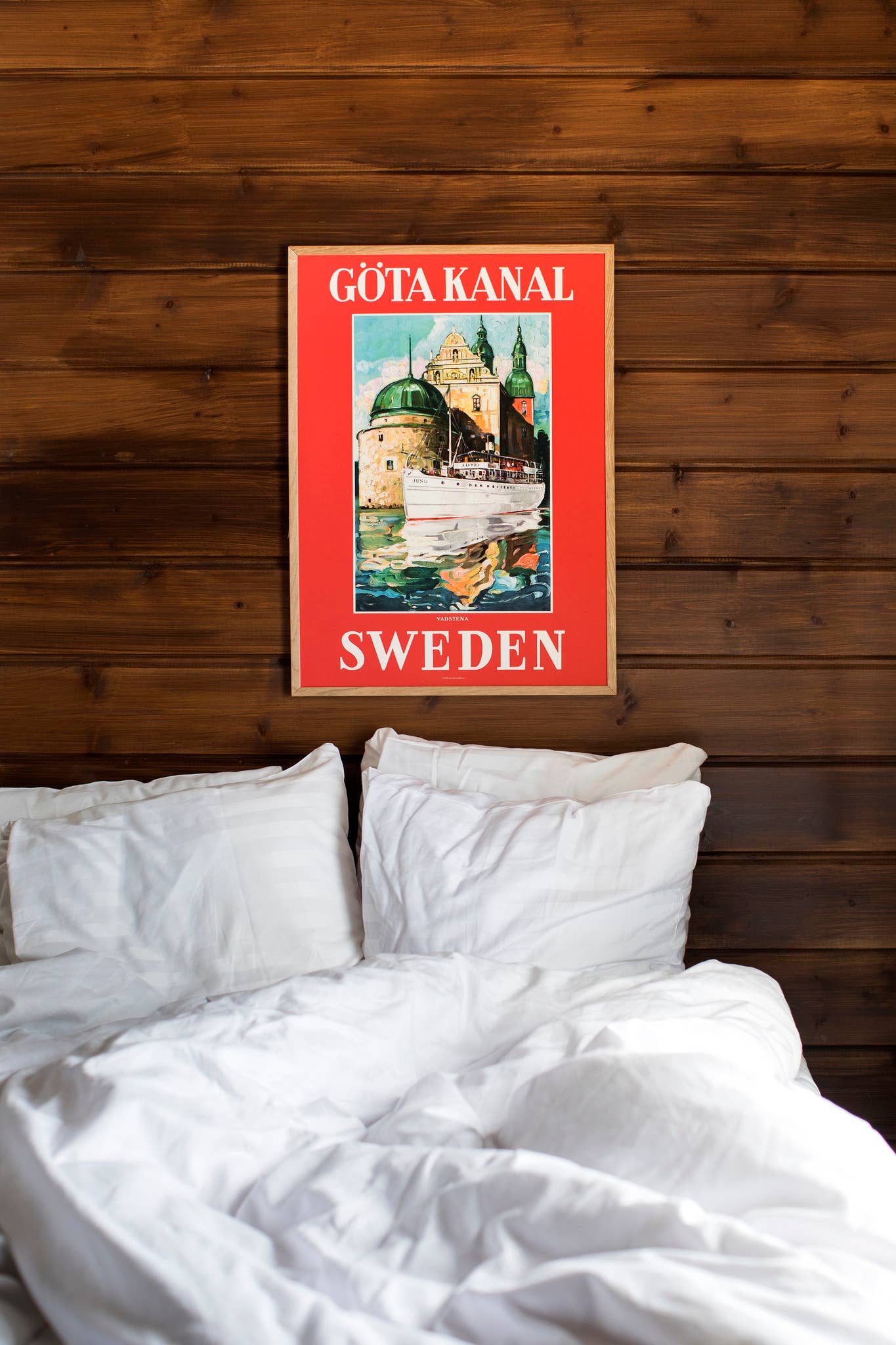 A wooden wall displays a 50 x 70 cm Scandinavia Travel Poster Göta Kanal Vadstena above an unmade white bed, printed on high-quality matte paper.
